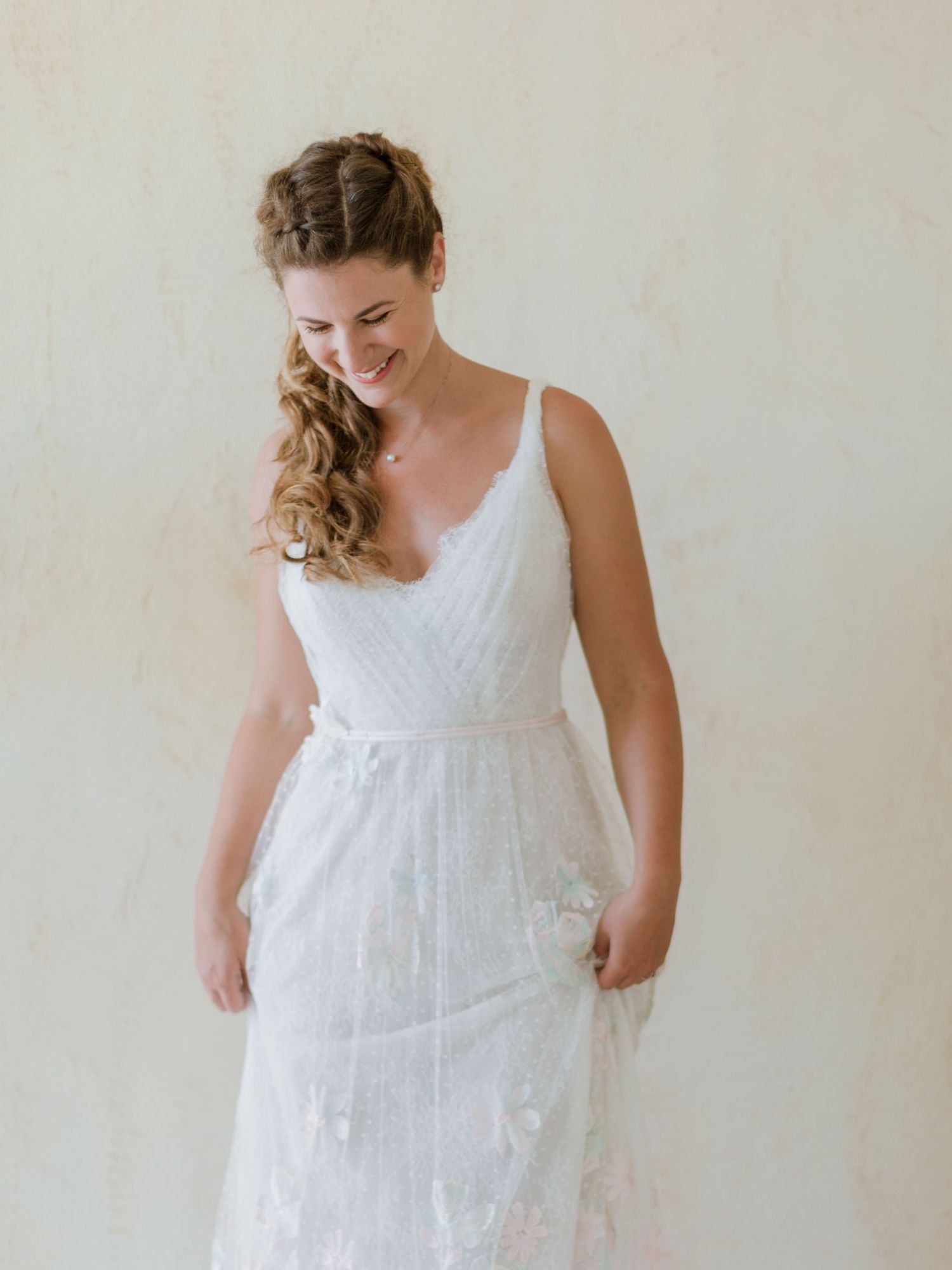 Mariée souriante portant une robe de mariée blanche en dentelle à fines bretelles avec des motifs floraux, et une coiffure en demi-queue