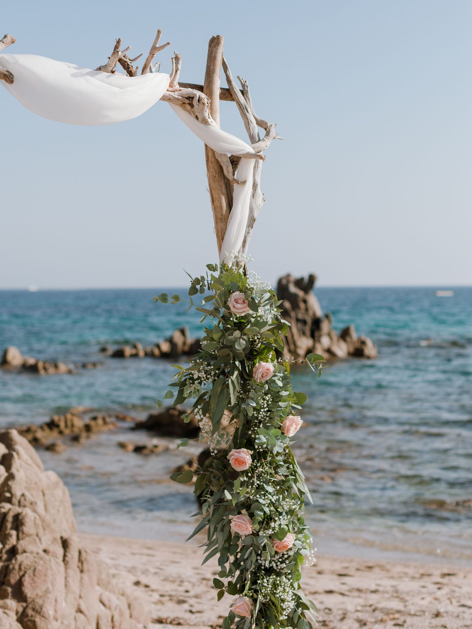 Arche de mariage en bois flotté ornée de roses roses et de feuillage, sur une plage rocheuse de la Méditerranée.