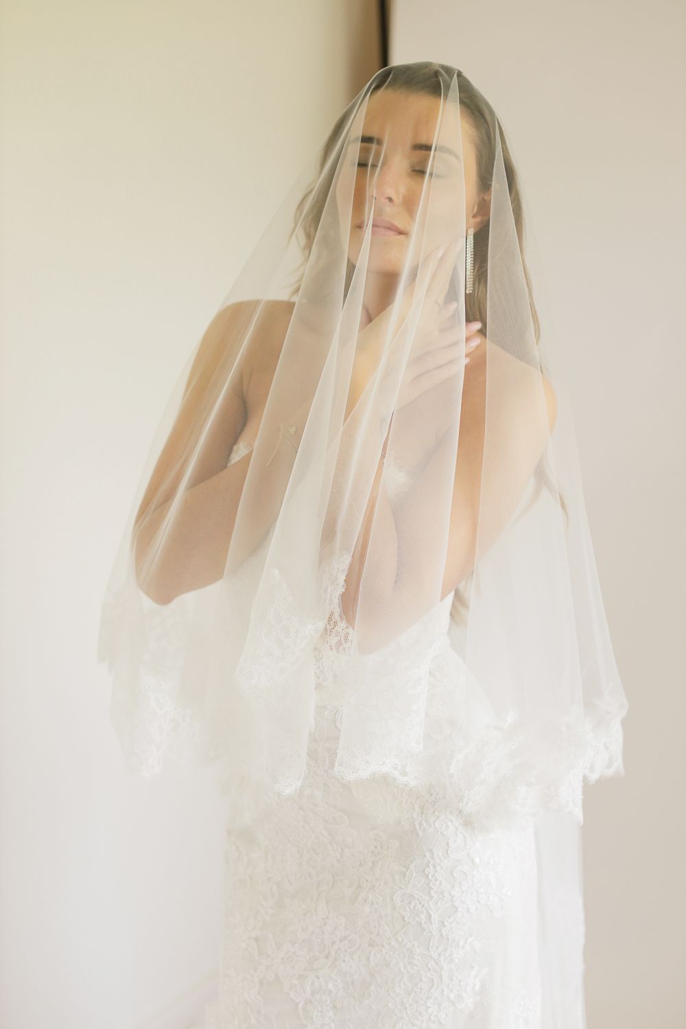 Bride in lace wedding dress wearing a delicate lace-trimmed veil draped over her face, posing elegantly.