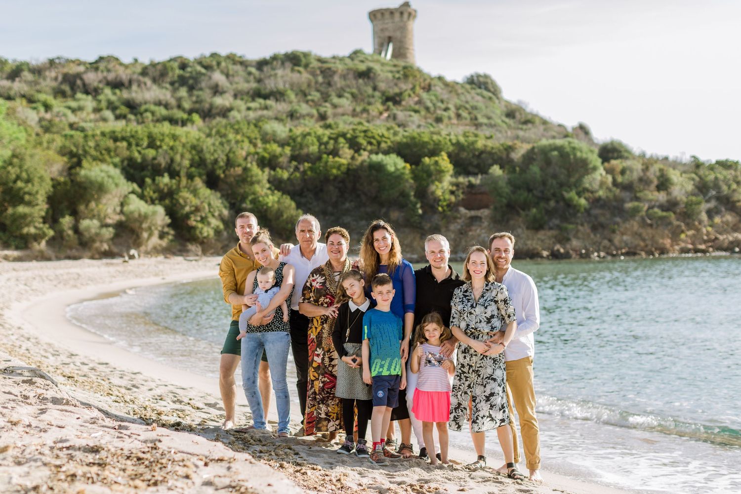 Groupe familial élargi posant sur une plage pittoresque avec une tour historique sur une colline en arrière-plan à Fautea, Corse.