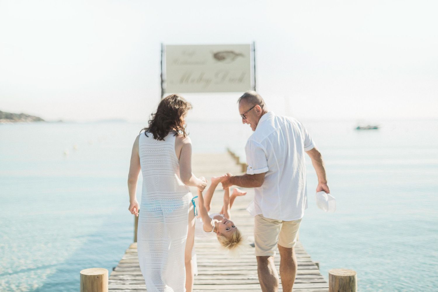Une famille joue sur une jetée en bois surplombant la mer bleue, les parents faisant balancer leur jeune enfant entre eux.