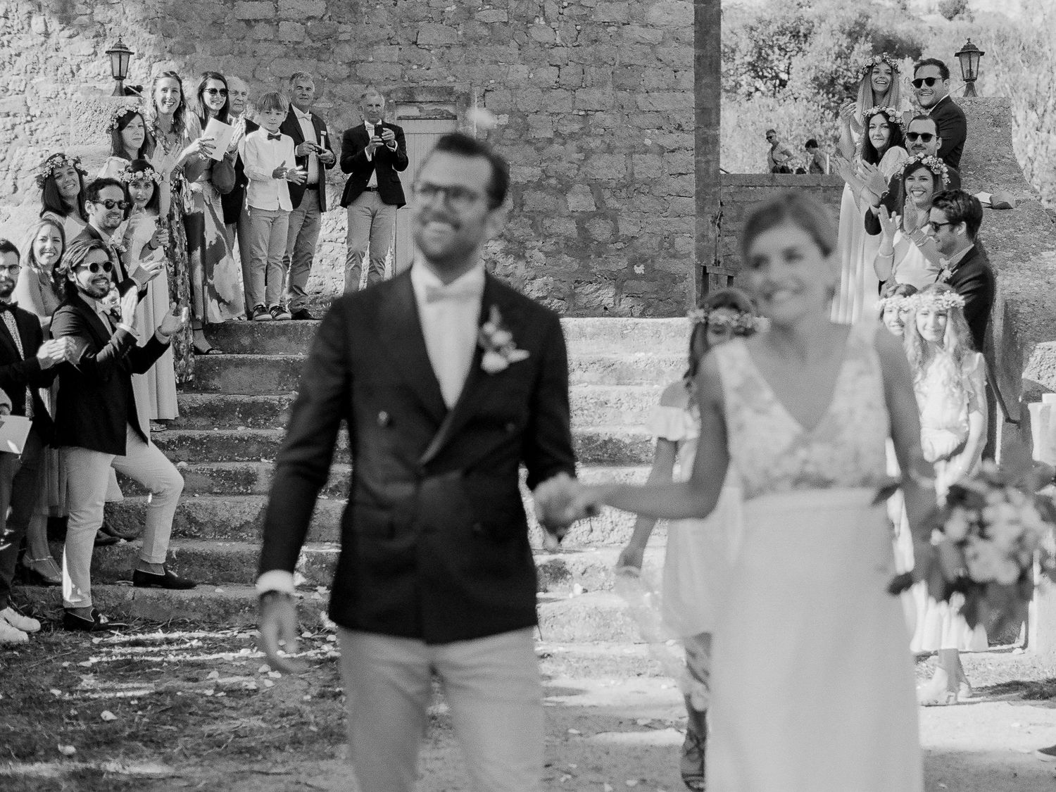 Photo de mariage en noir et blanc d'un couple marchant main dans la main devant des invités qui les applaudissent sur des marches en pierre.