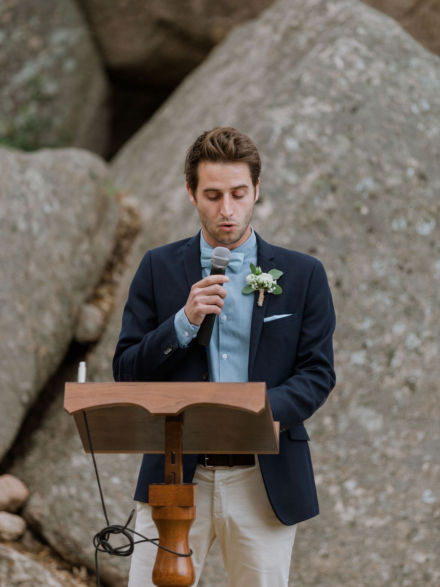 Un jeune homme en blazer bleu marine et nœud papillon prend la parole à un podium en bois lors d'une cérémonie de mariage en plein air.