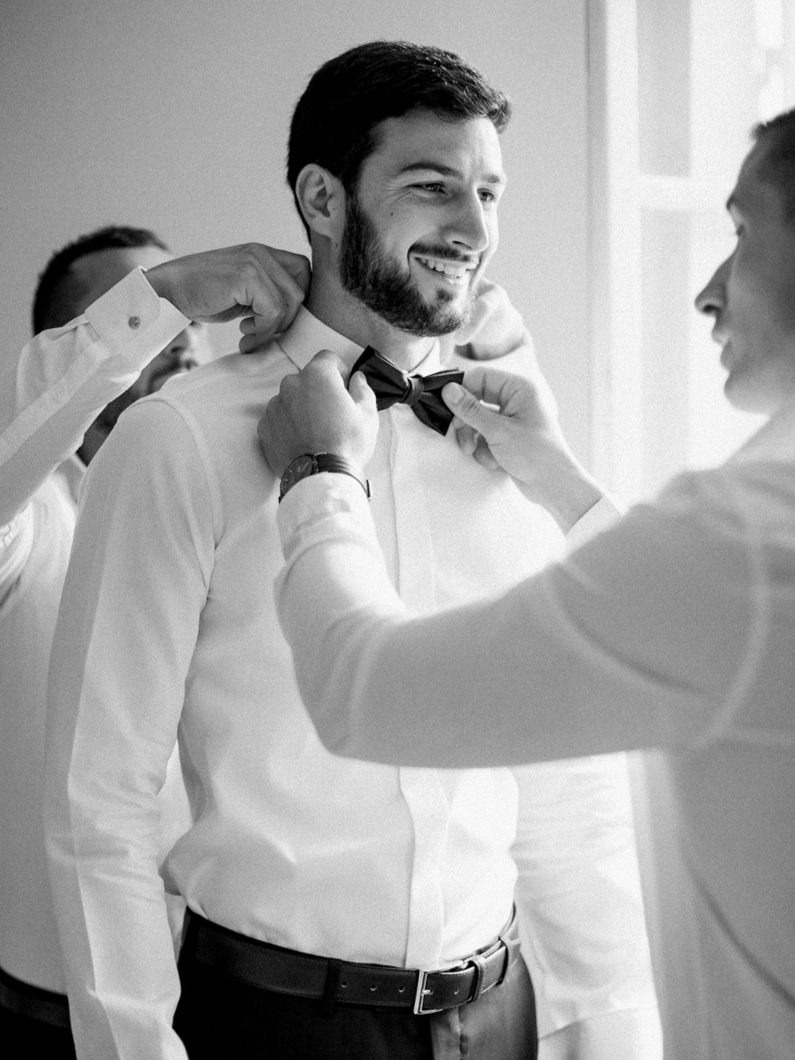 Black and white photo of a smiling groom getting his bow tie adjusted by groomsmen before his wedding ceremony.