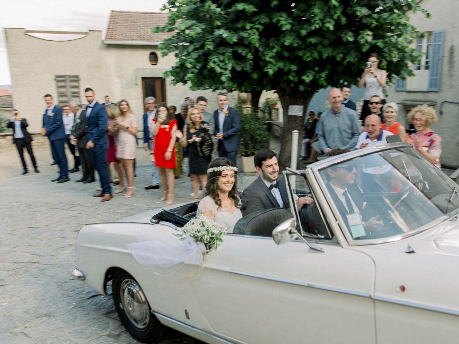 Newlyweds ride in a classic white convertible car surrounded by wedding guests in a charming village square.