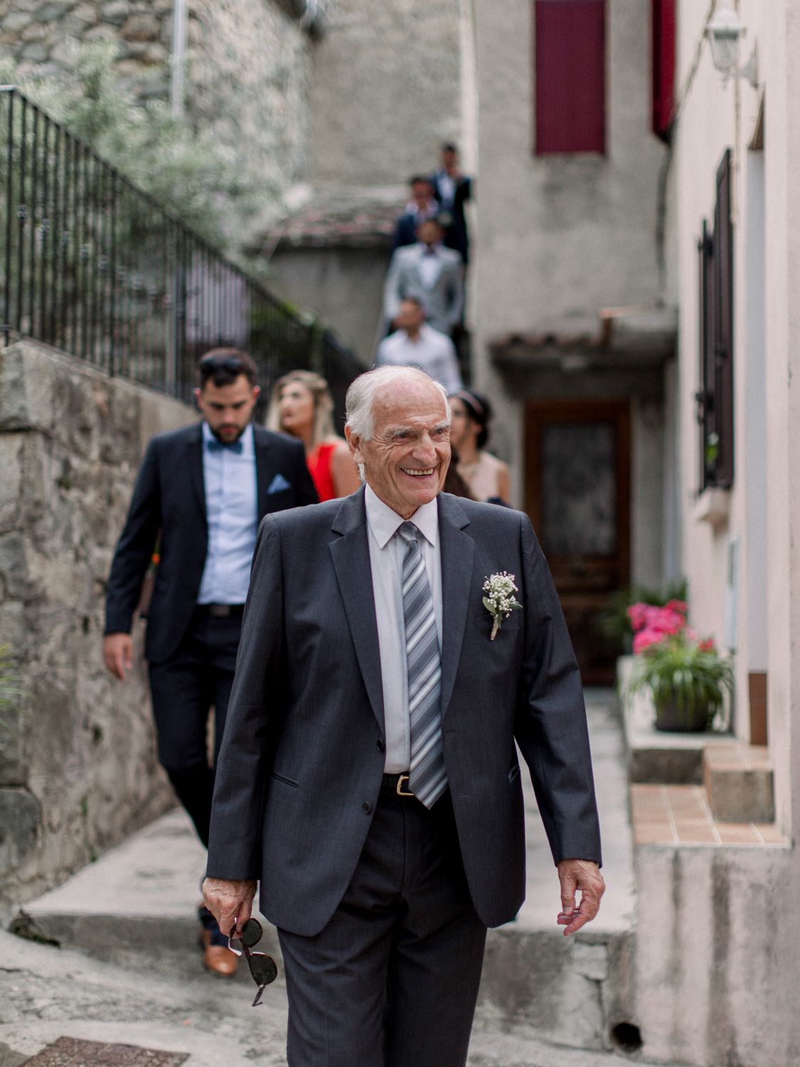 Smiling older man in suit with boutonniere leads wedding party down stone steps in European village.