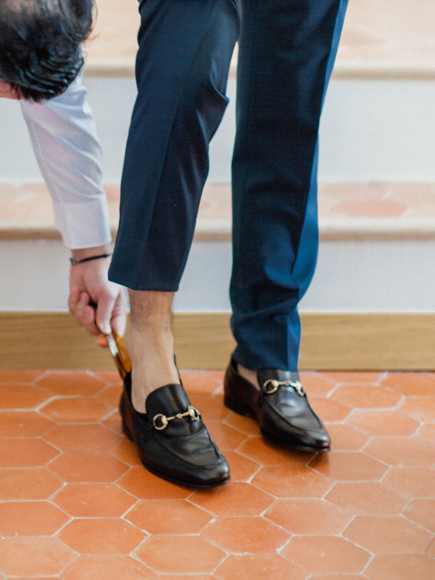 Man wearing navy trousers putting on black leather Gucci horsebit loafers on terracotta tile floor.