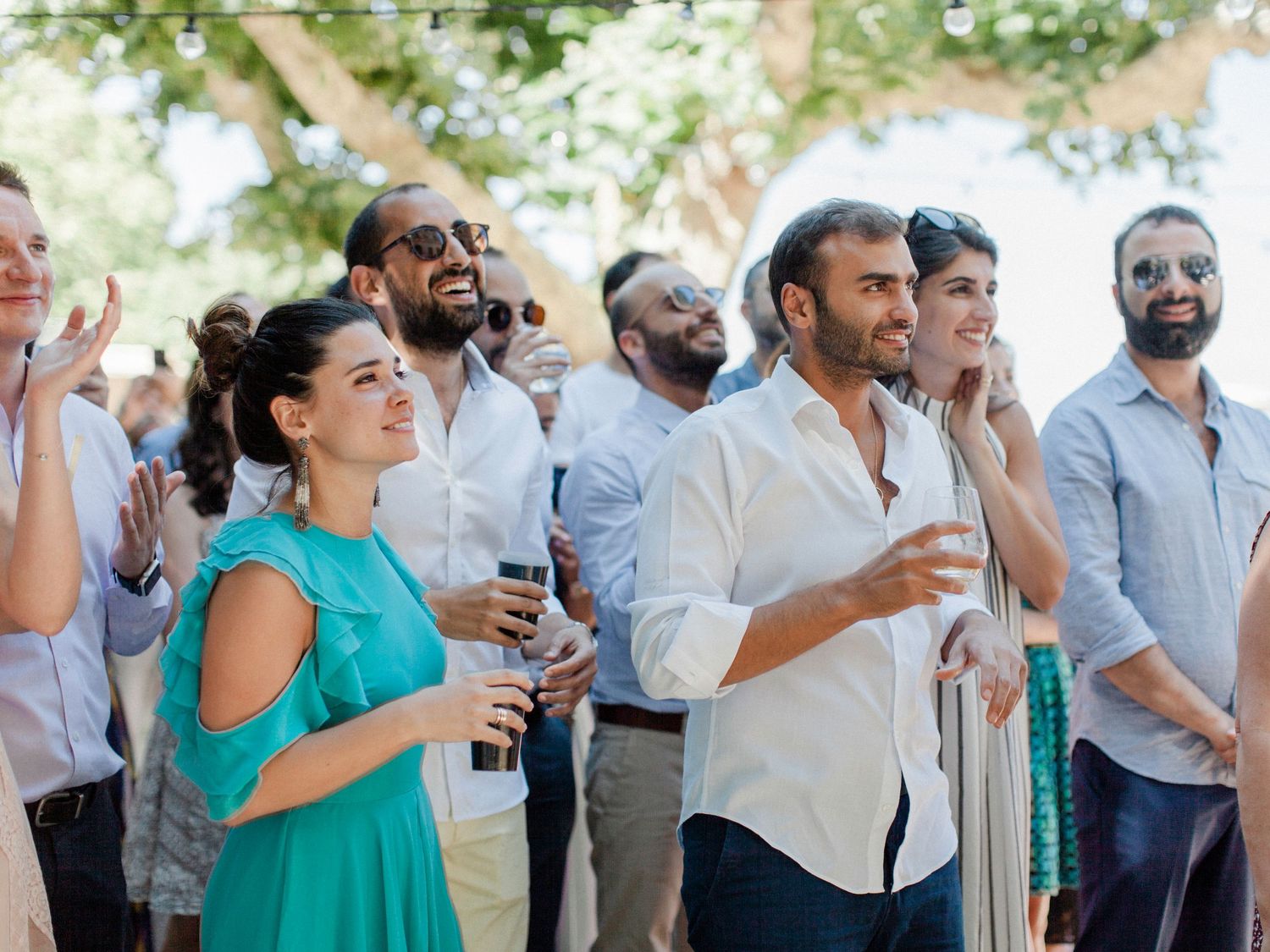 Group of elegantly dressed guests laughing and clapping at an outdoor wedding reception on a sunny day.