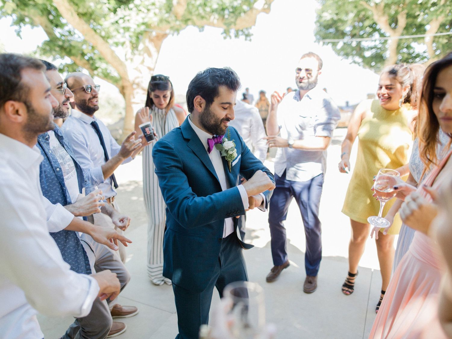 Groom in navy suit celebrates with wedding guests outdoors, clapping and dancing under trees on a sunny day.