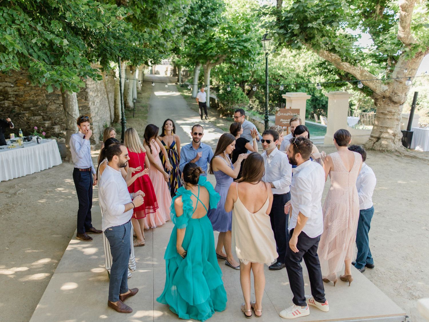 Guests dancing and socializing outdoors at a wedding reception under shaded trees on a sunny day.