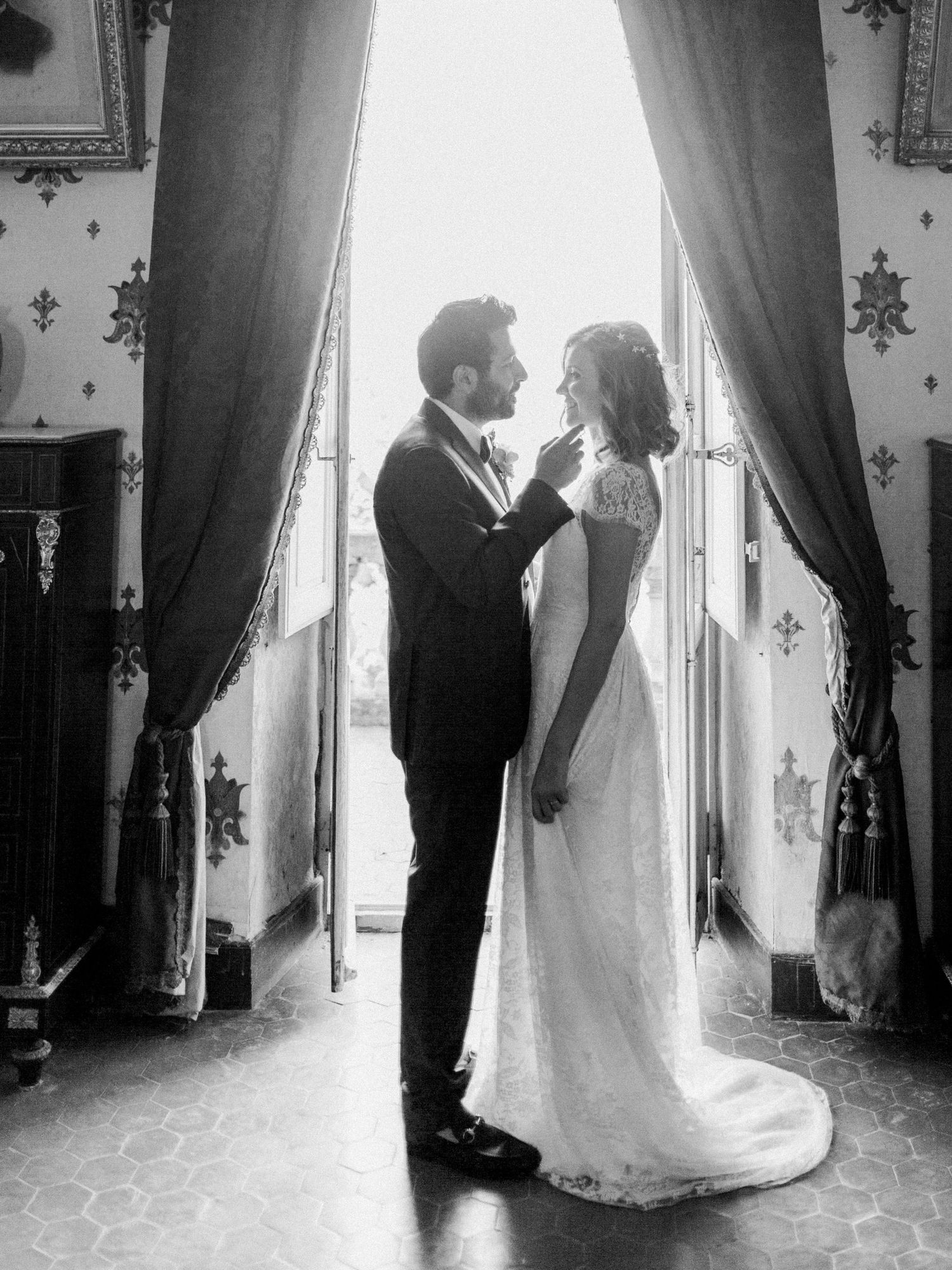 Black and white photo of a bride and groom sharing an intimate moment in an elegant room with draped curtains.