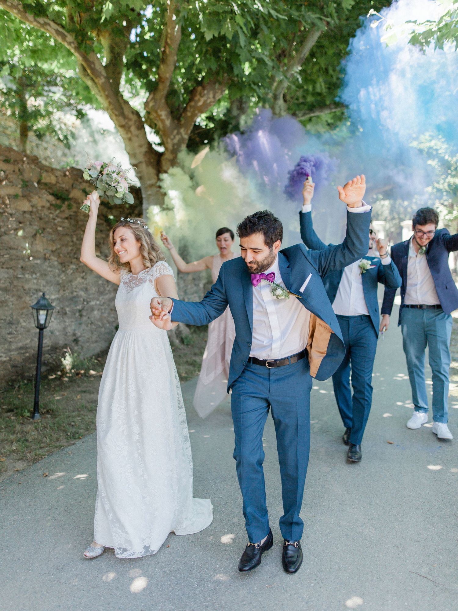 Bride and groom celebrate with colorful purple smoke bombs, flanked by groomsmen in blue suits outdoors.