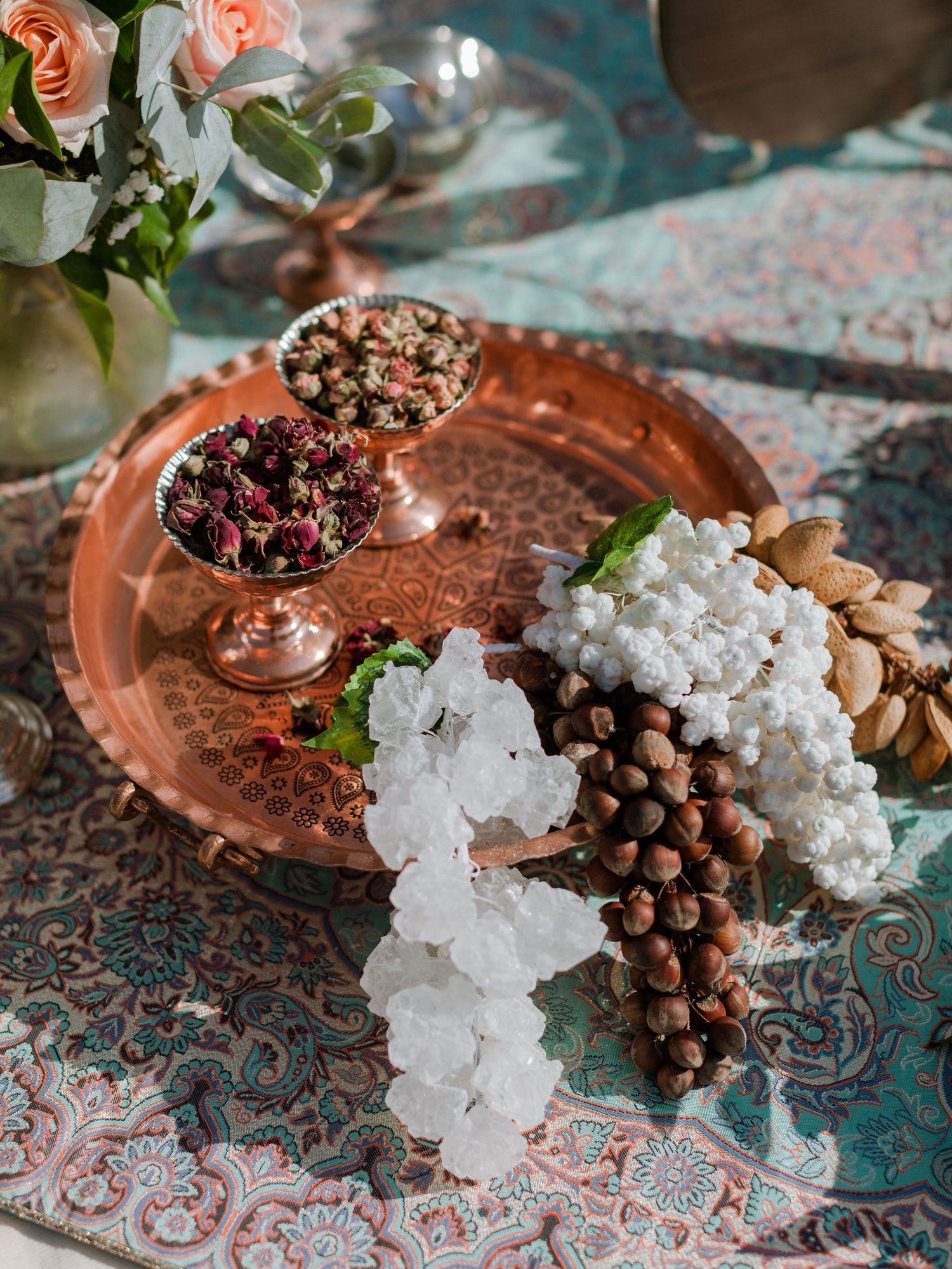 Plateau en cuivre avec des boutons de rose, des noix et des cristaux de sucre blanc, posé sur une nappe à motifs cachemire et fleurs roses.