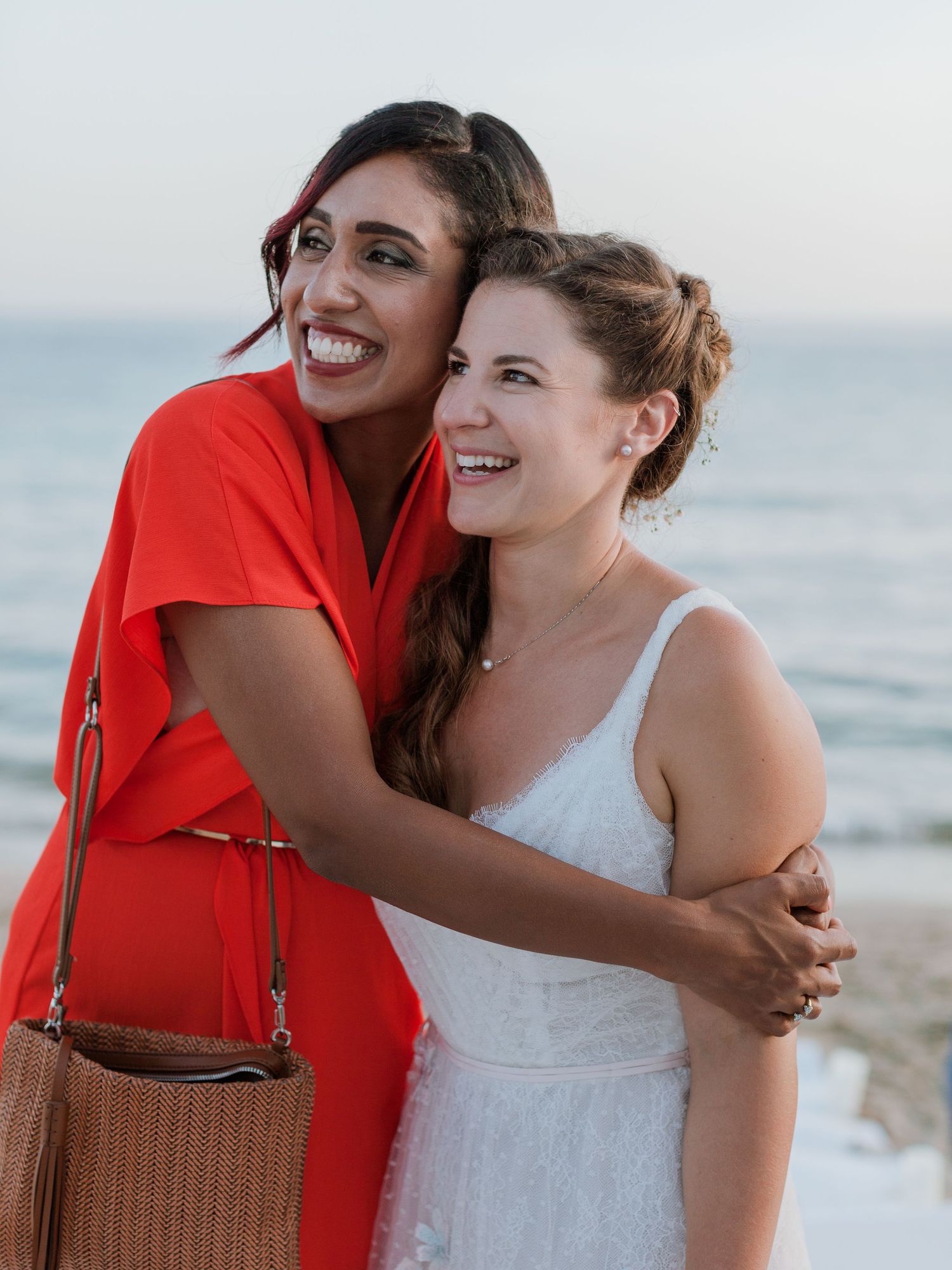 Deux femmes souriantes s'étreignant sur une plage au coucher du soleil, l'une portant une combinaison rouge et l'autre une robe blanche.