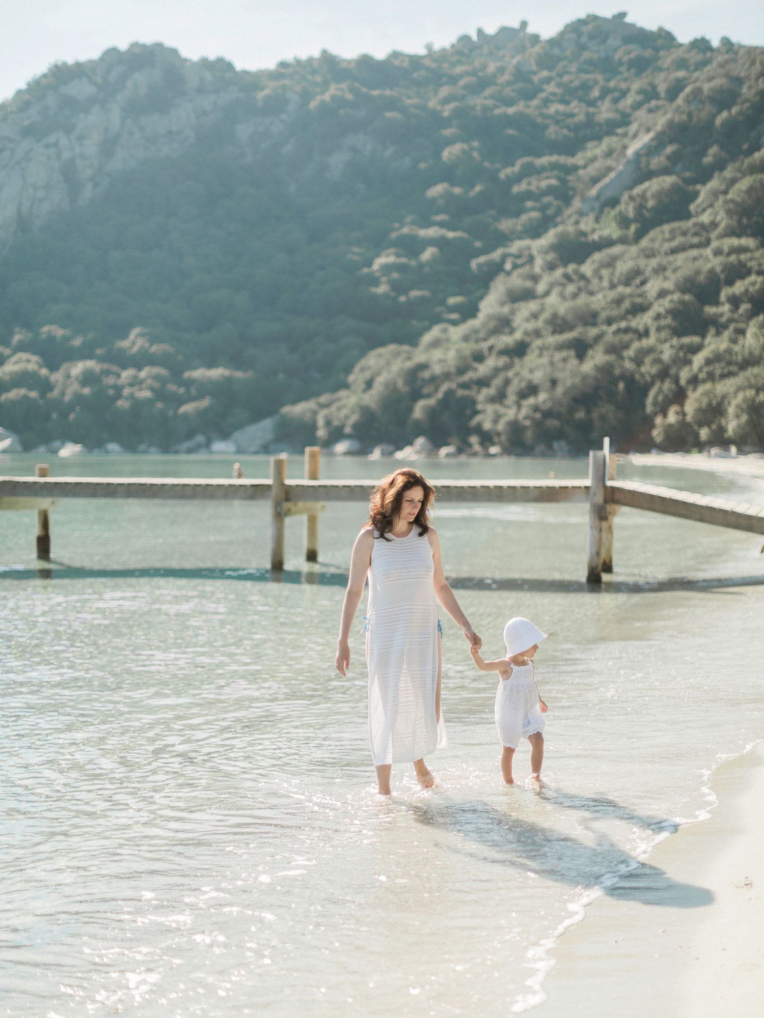 Une mère en robe blanche marche main dans la main avec son jeune enfant le long d'une plage  ensoleillée à Santa Giulia corse.