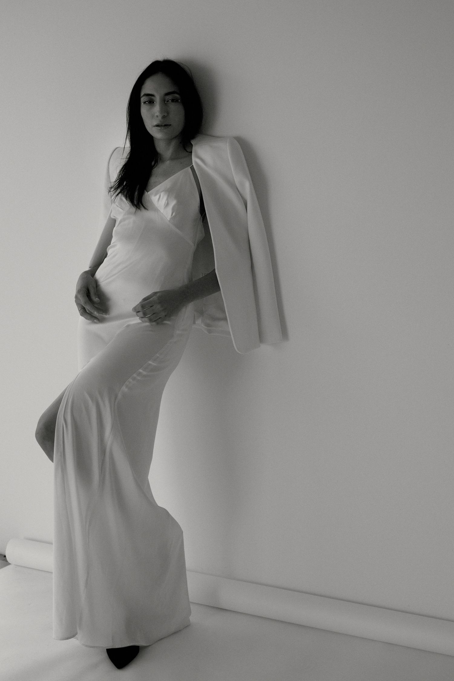 Photo en noir et blanc d'une femme portant un pantalon blanc évasé et un blazer posé sur l'épaule, prenant la pose devant un mur blanc.