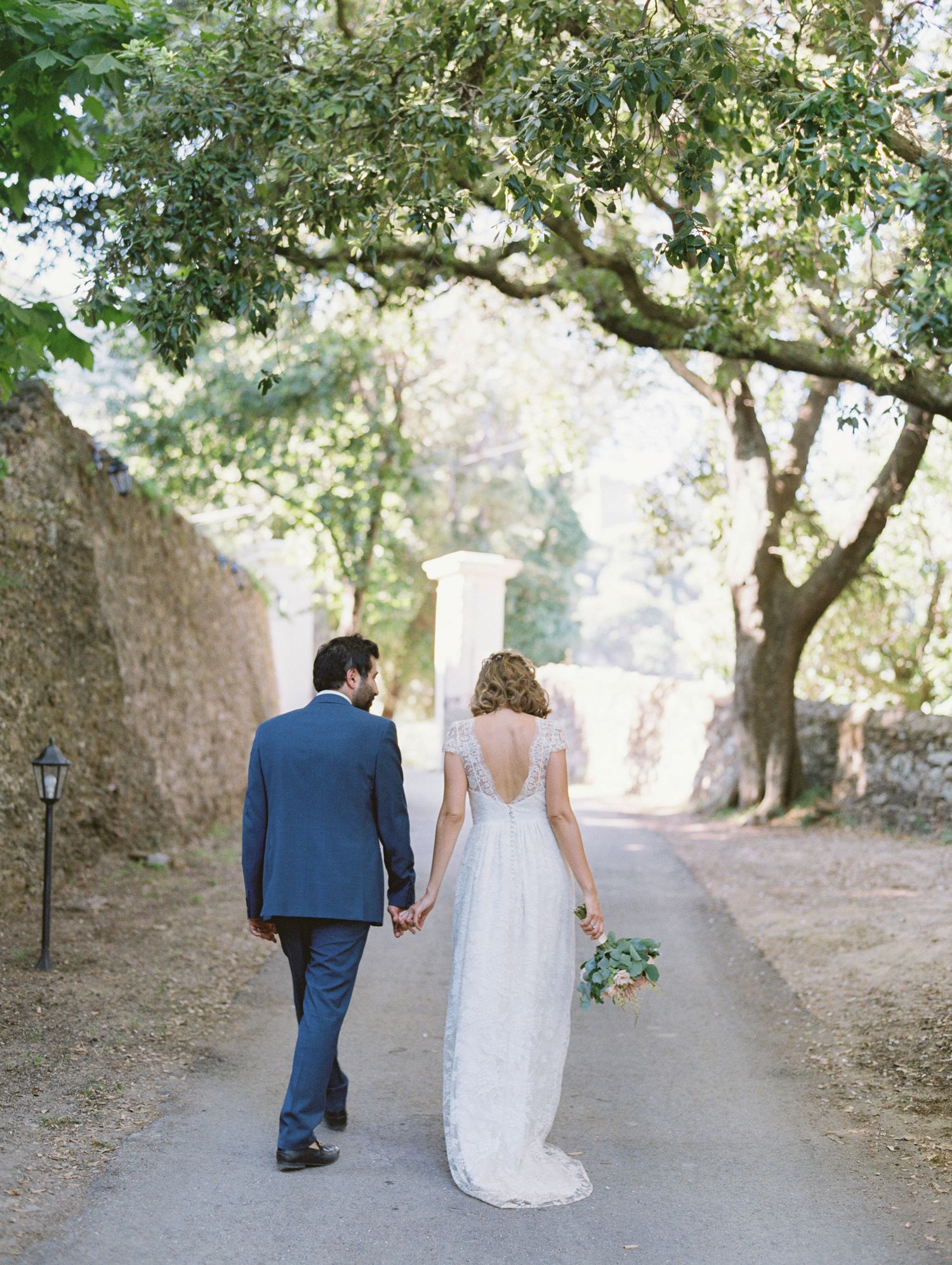 Bride and groom walking hand in hand down a tree-lined path, bride in lace gown holding bouquet.