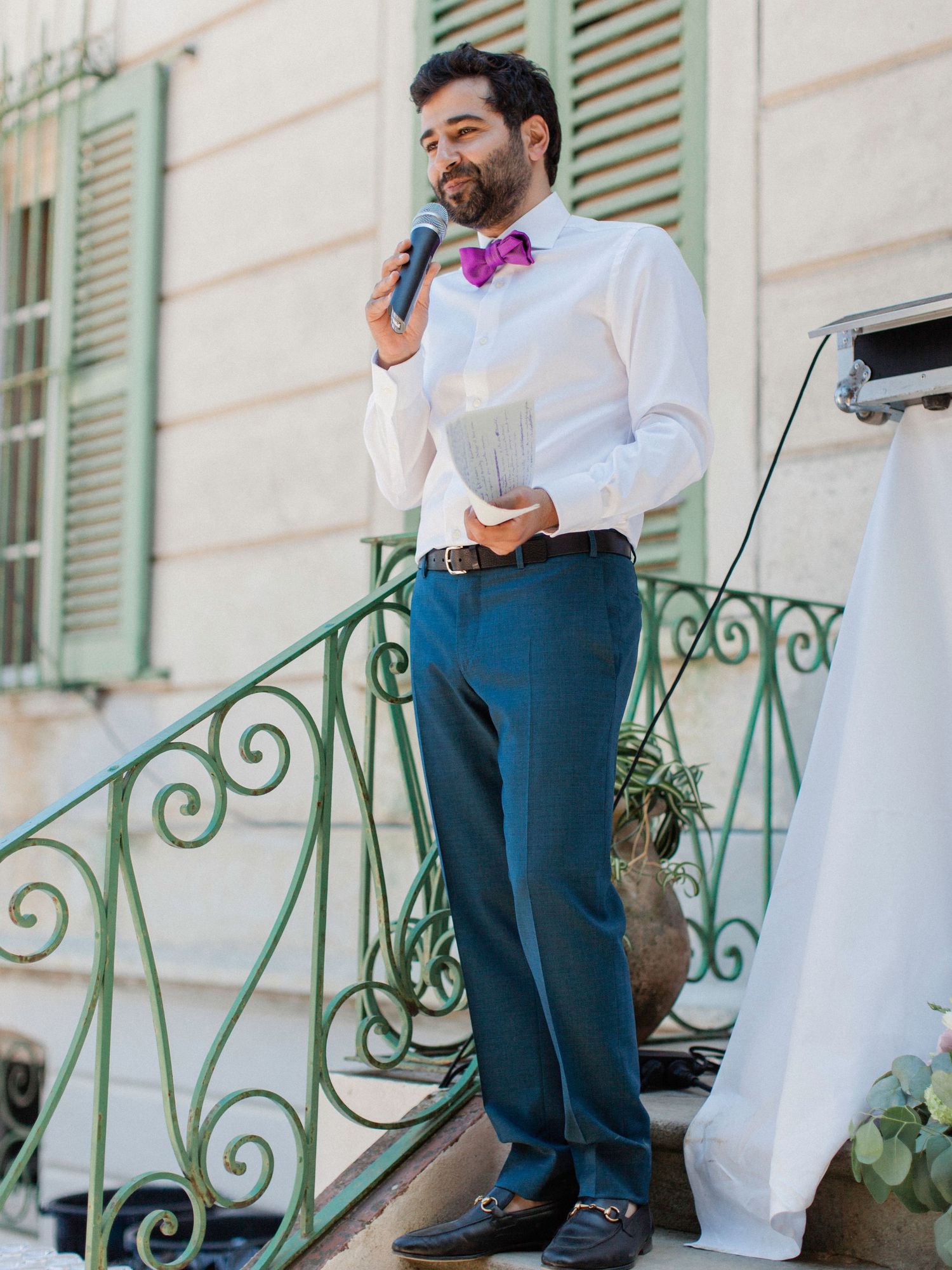 Man in white shirt and bow tie speaks into microphone at outdoor wedding reception on decorative steps.