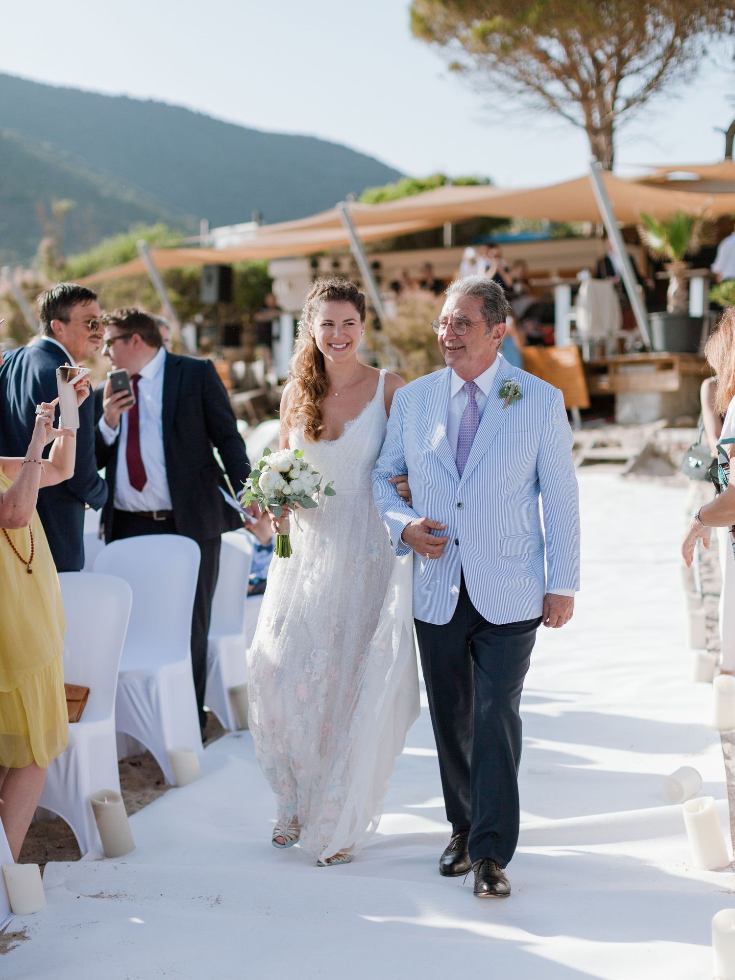La mariée descend l'allée extérieure au bras de son escorte lors d'une cérémonie de mariage sur la plage, tenant un bouquet blanc