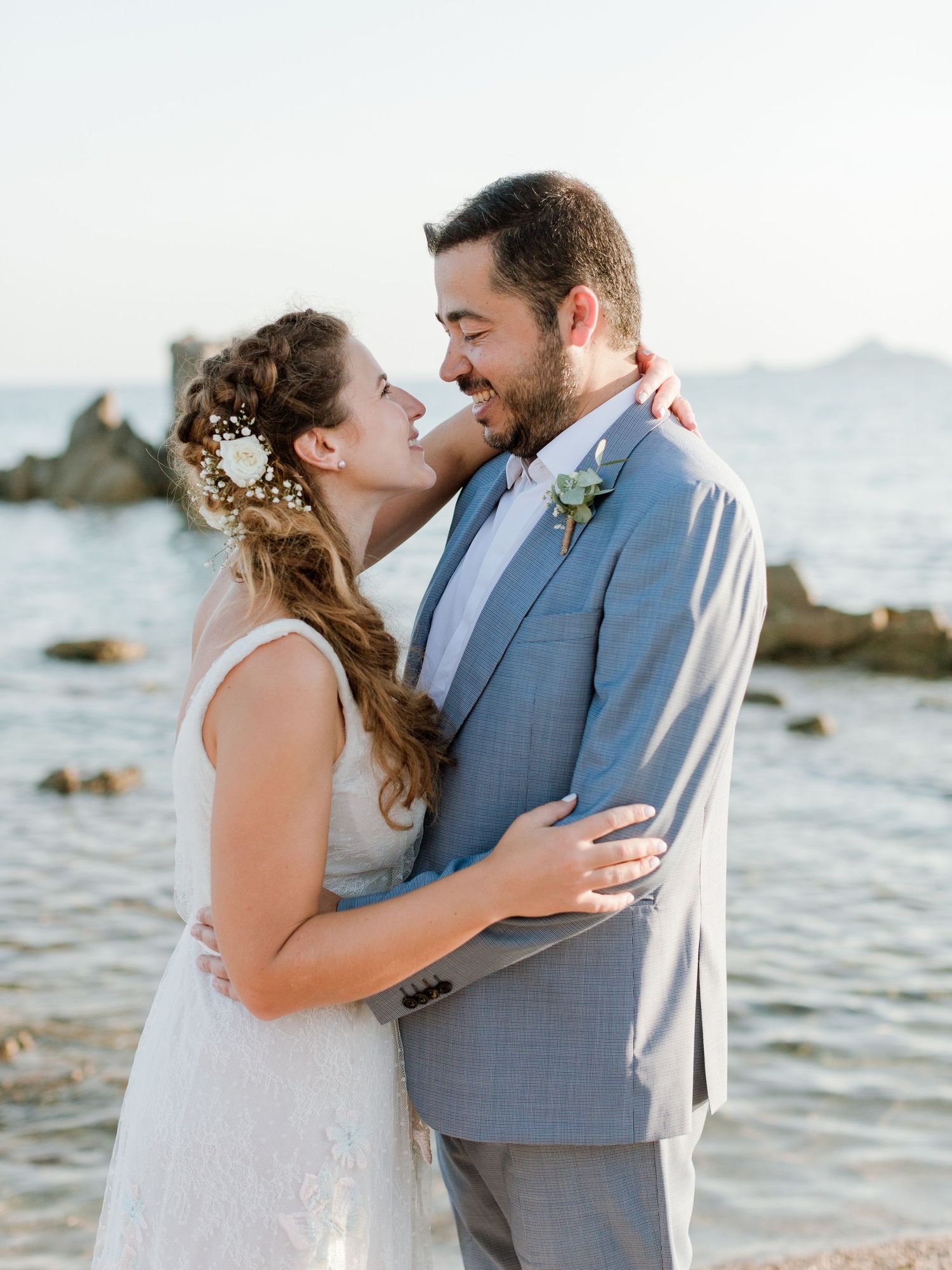 Les mariés partagent un moment de joie en s'étreignant sur une plage de galets ; elle porte une robe blanche et un accessoire floral dans le
