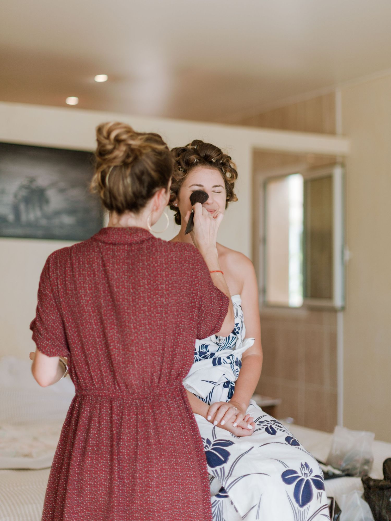 Une maquilleuse en robe rouge maquille une mariée portant des bigoudis dans une chambre lumineuse.