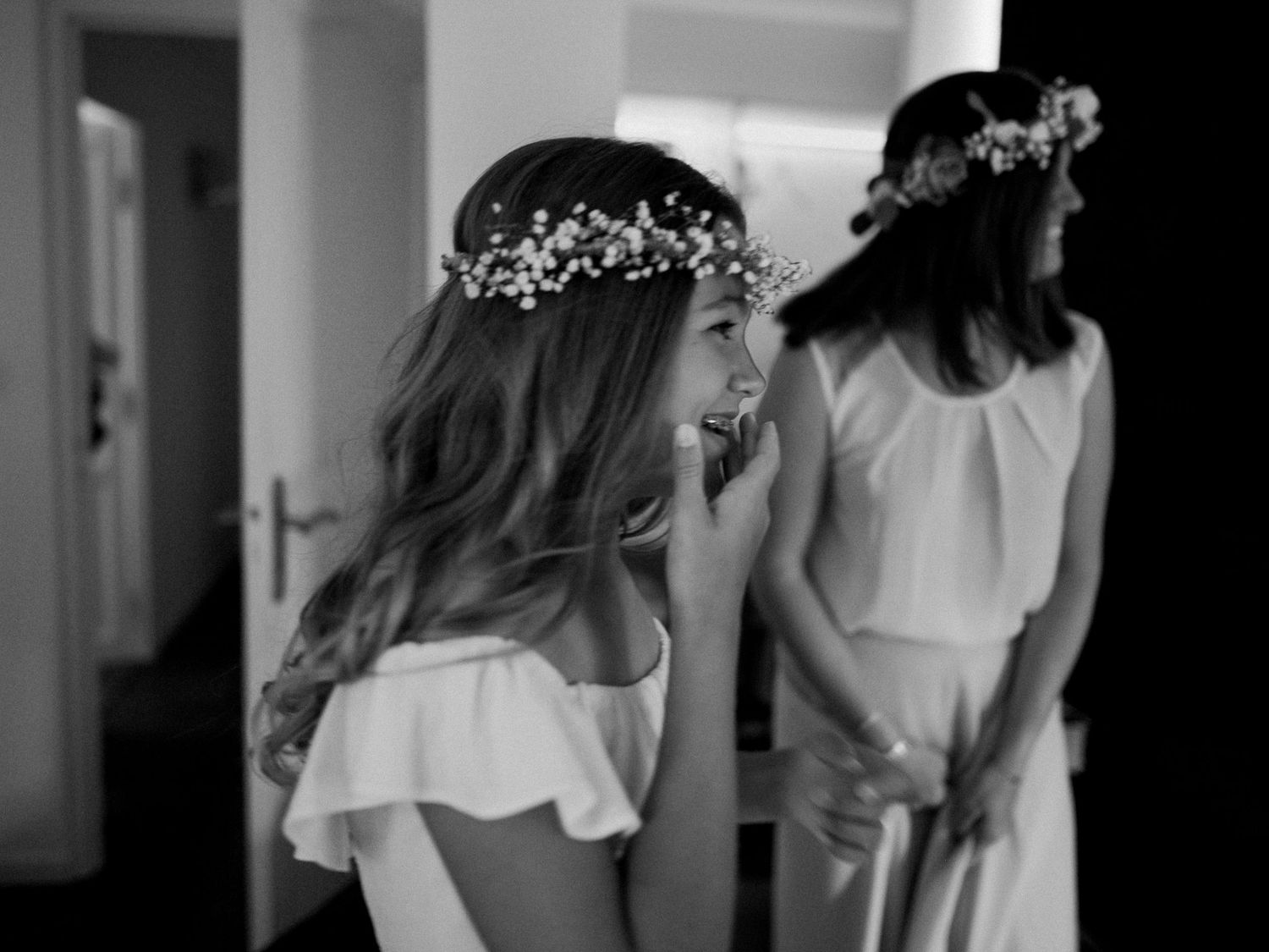 Deux jeunes filles d'honneur en robes blanches et couronnes de fleurs rient ensemble sur une photo de mariage en noir et blanc.