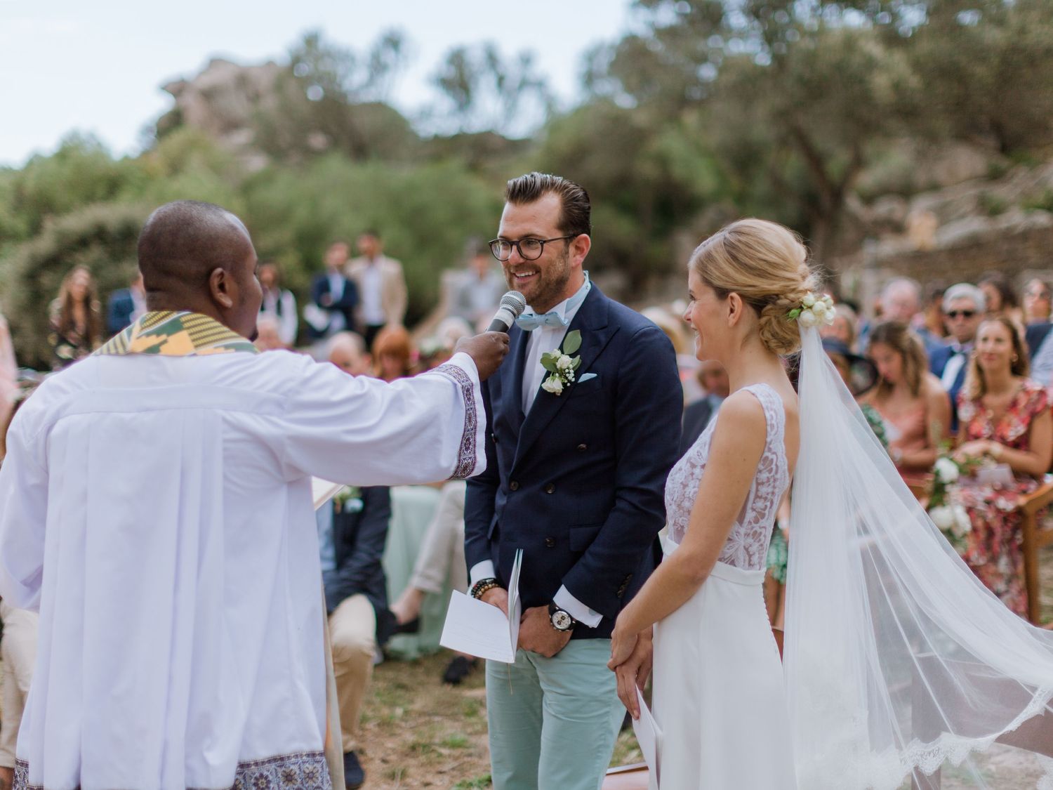 Cérémonie de mariage en plein air : le marié porte un costume bleu marine, la mariée un voile blanc, et l'officiant bénit les époux.
