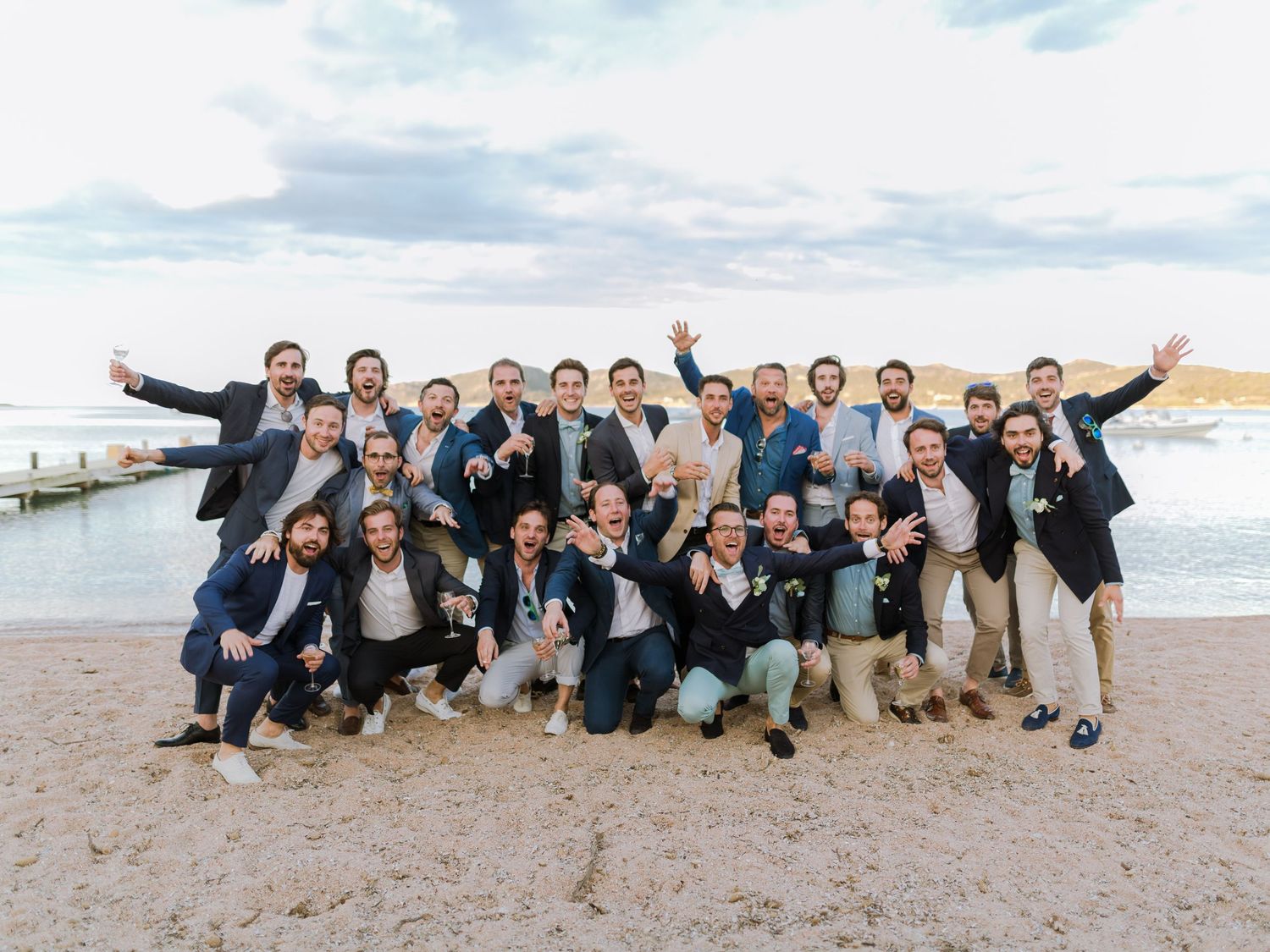 Un grand groupe d'hommes en tenue de cérémonie pose ensemble sur une plage de sable fin lors d'une réception de mariage en bord de mer.