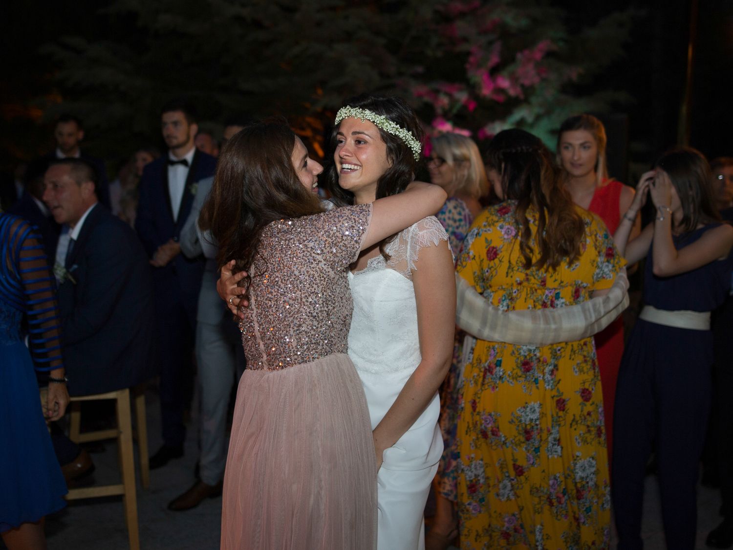 La mariée en robe blanche et couronne de fleurs enlace une invitée vêtue d'une robe à paillettes