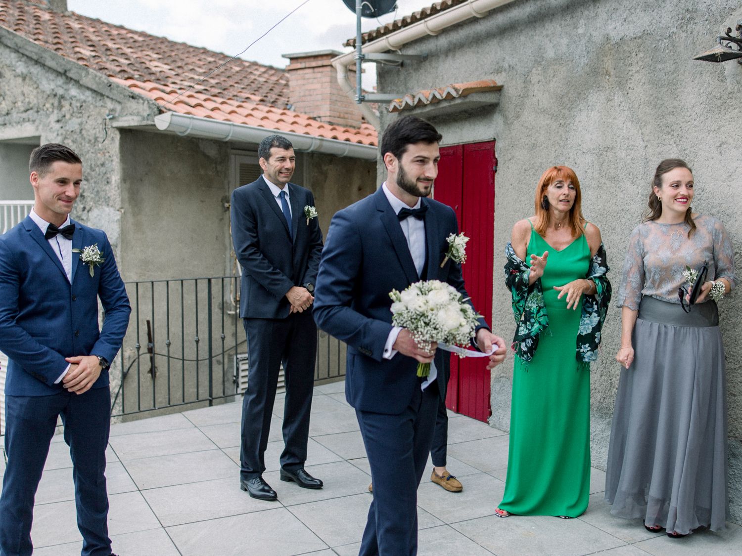 Groom in navy suit holds bouquet outdoors at wedding ceremony with guests in formal attire near stone building.