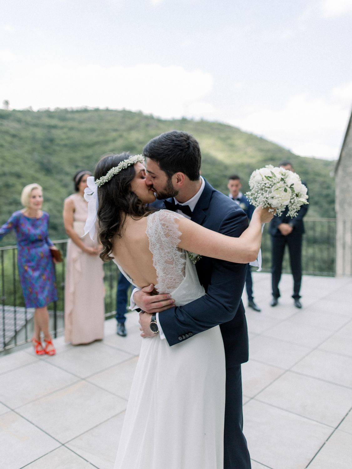 Bride and groom share first kiss on terrace with wedding guests and green hills in background.