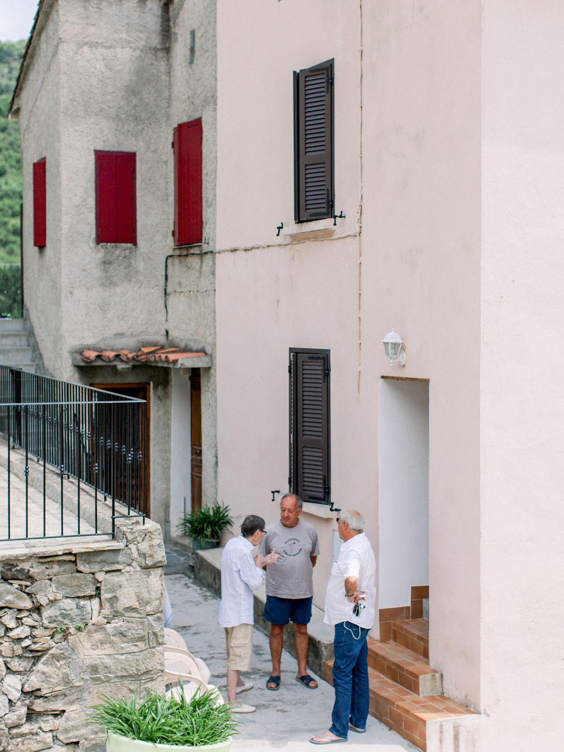 Three people conversing outside a rustic European stone building with red and dark shuttered windows.
