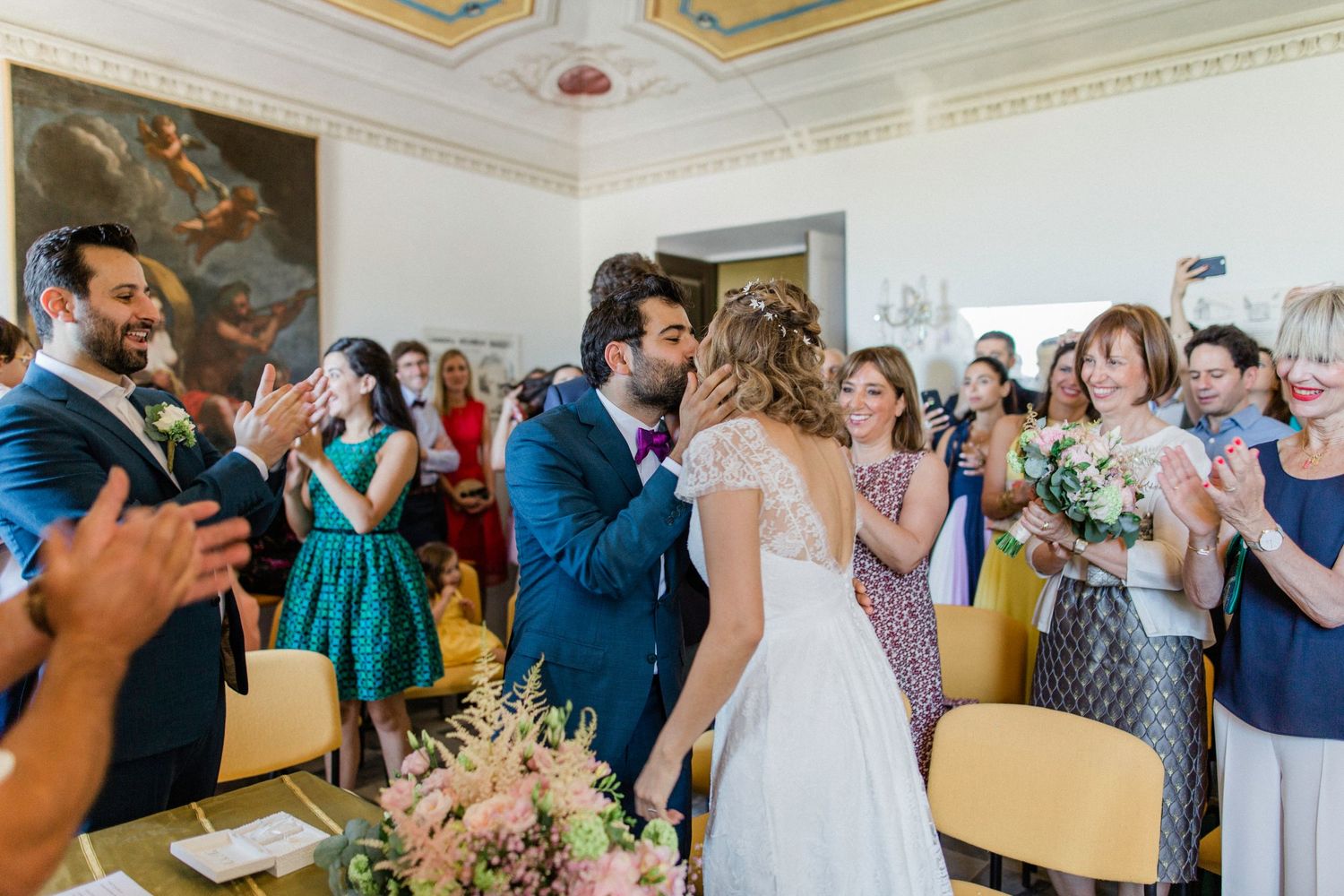 Bride and groom share a first kiss surrounded by applauding guests at an elegant civil wedding ceremony.