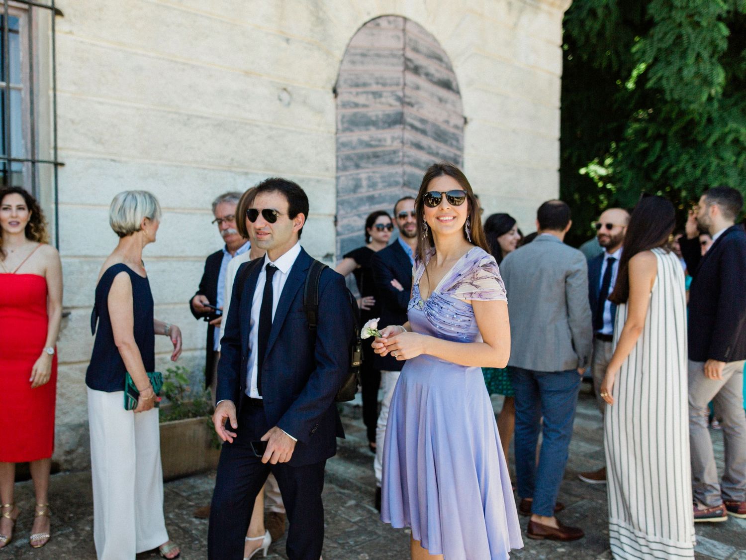 Elegantly dressed guests mingle outdoors at a summer wedding reception, with a man in a suit and woman in lavender dress.
