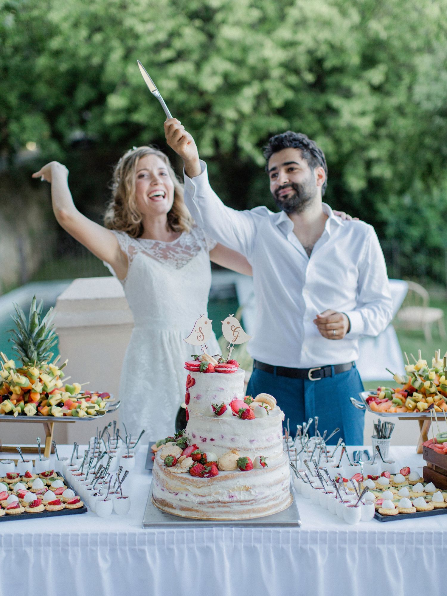 Happy couple cutting a three-tier naked wedding cake decorated with strawberries at an outdoor dessert table.