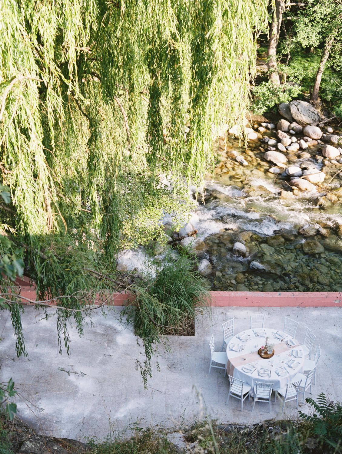 Aerial view of a white dining table beside a rocky stream, shaded by weeping willow trees on a stone terrace.