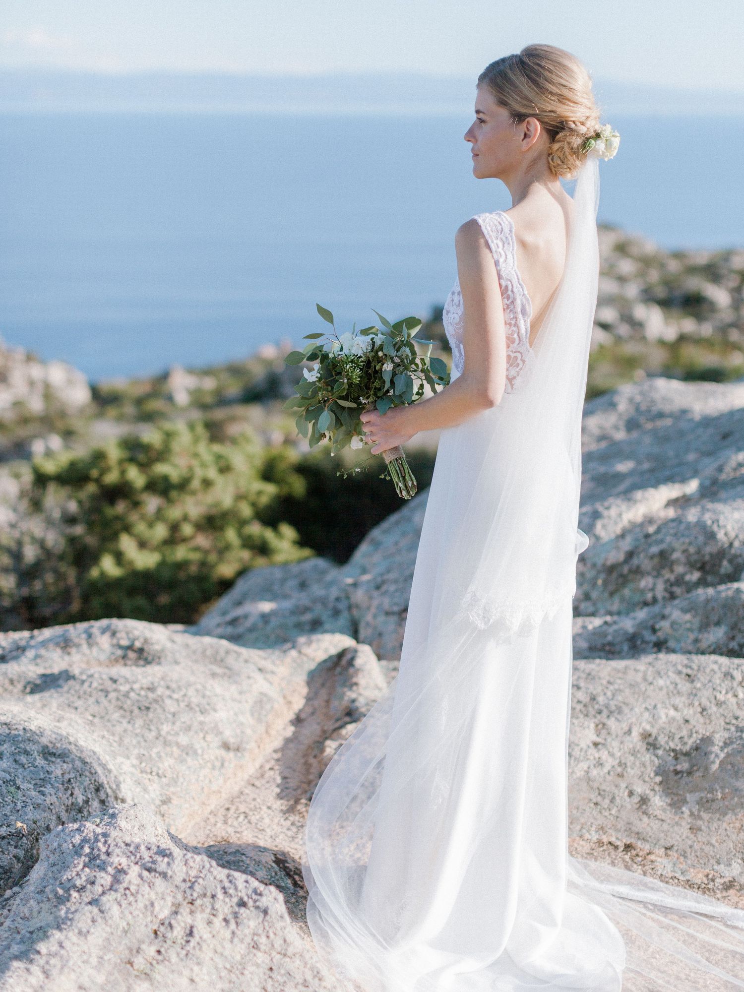 Une mariée en robe de dentelle blanche tient un bouquet de verdure sur une falaise rocheuse côtière avec vue sur la mer à Bonifacio