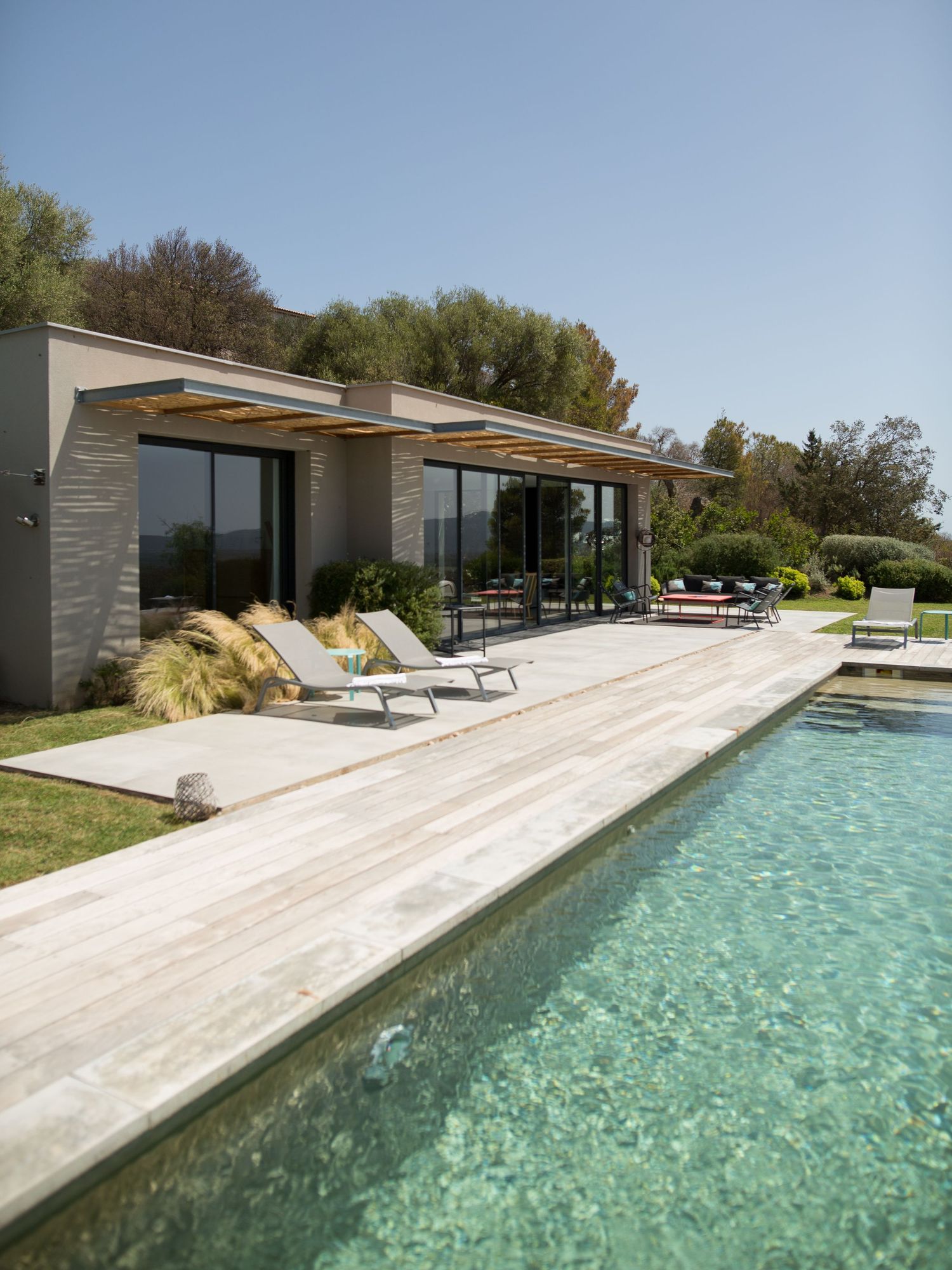 Villa moderne avec grande piscine, terrasse en bois et chaises longues sous un ciel bleu azur à Porto-Vecchio.