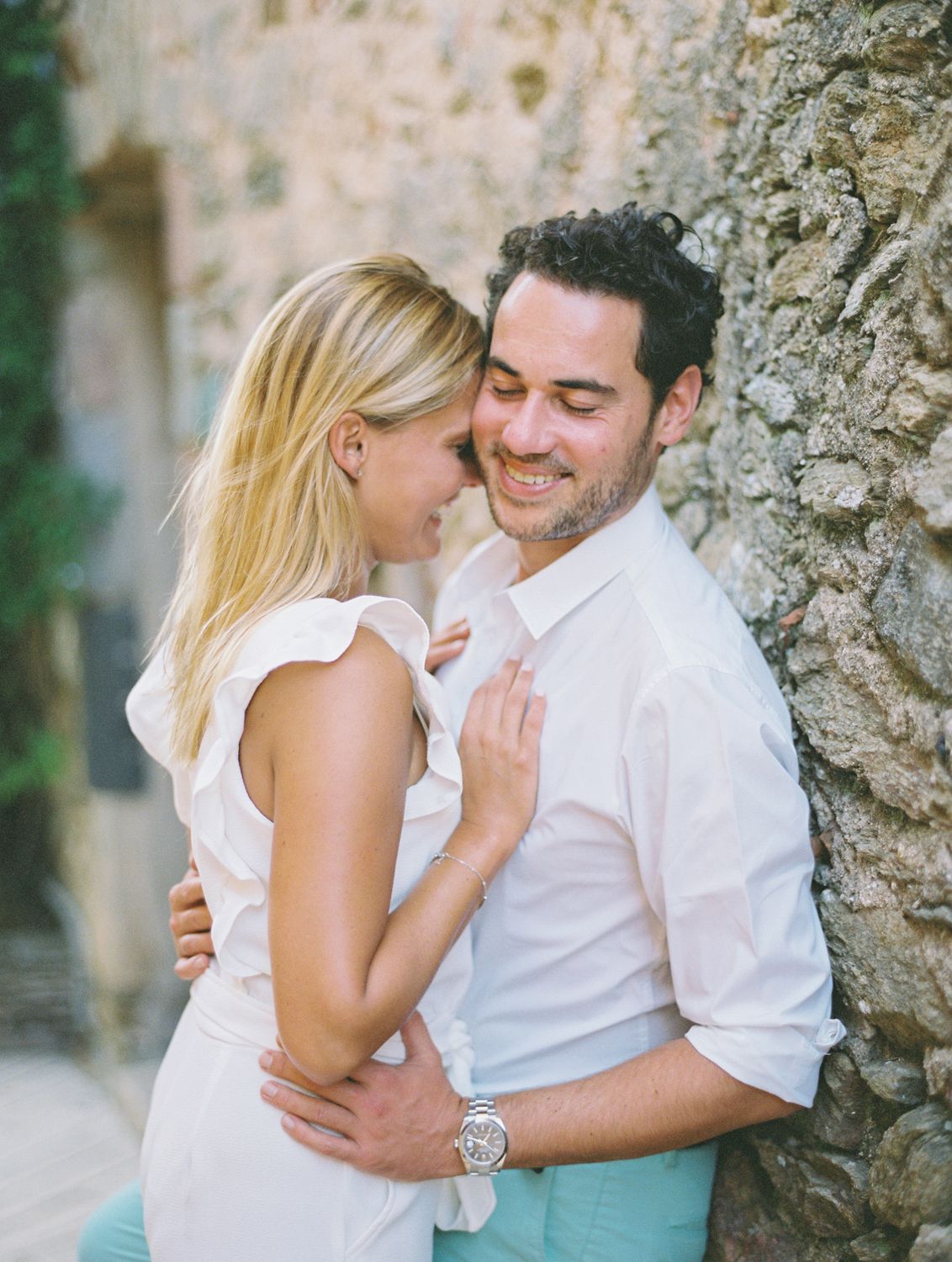 Un couple souriant, vêtu de blanc, s'enlace tendrement devant un mur de pierre rustique, à l'extérieur. A Gassin en Provence