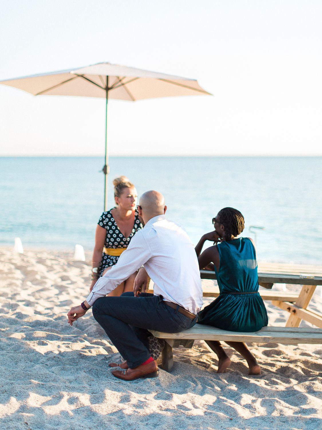 Trois personnes sont assises à une table de pique-nique sur la plage, sous un parasol blanc, en Corse.