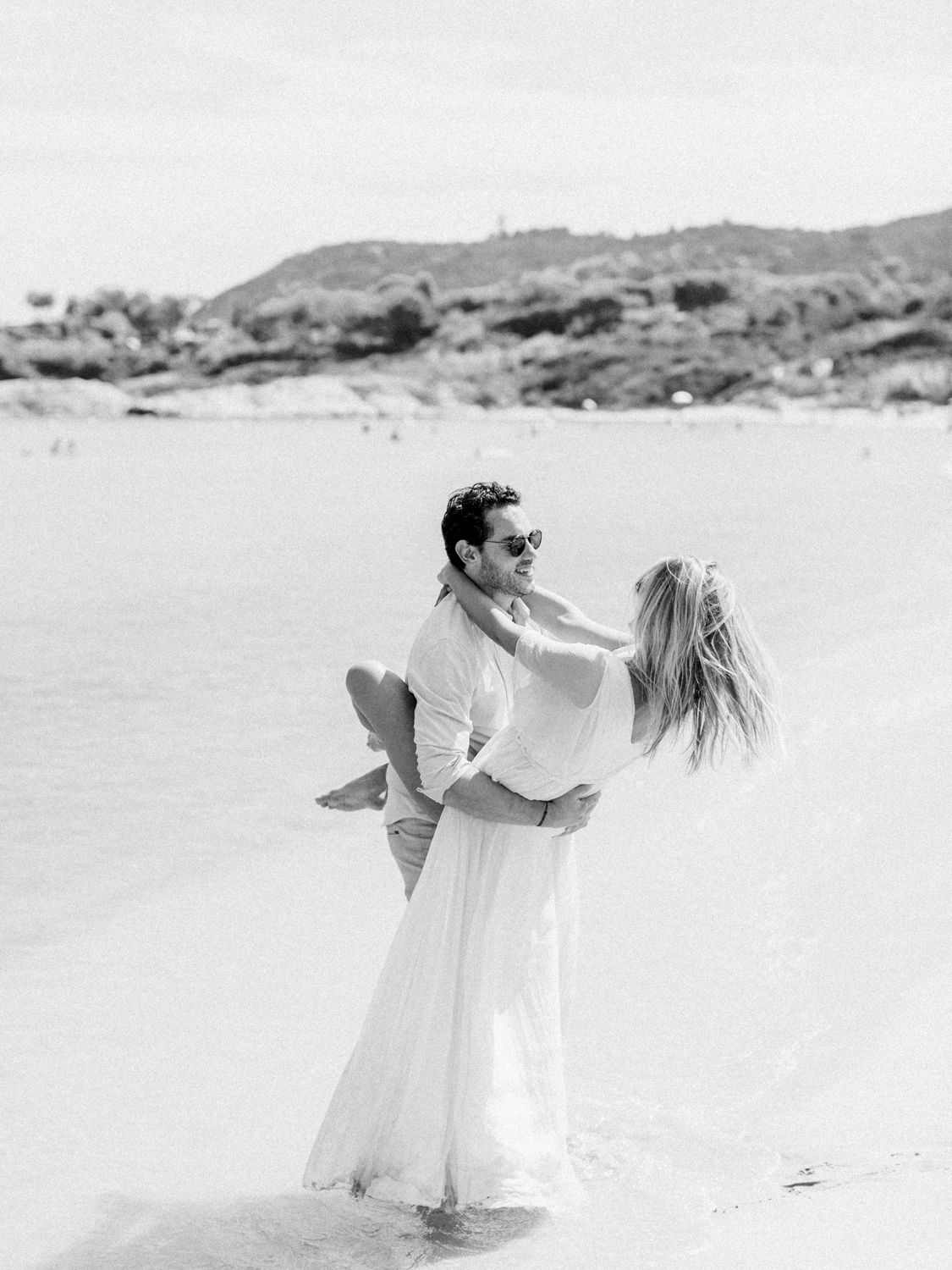 Photo en noir et blanc d'un couple enlacé sur une plage, femme en robe blanche, homme portant des lunettes de soleil, eau peu profonde.