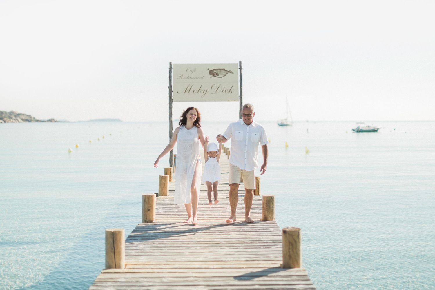 Une famille heureuse, vêtue de blanc, se promène sur une jetée en bois au-dessus d'une eau turquoise cristalline. Plage de Santa Giulia