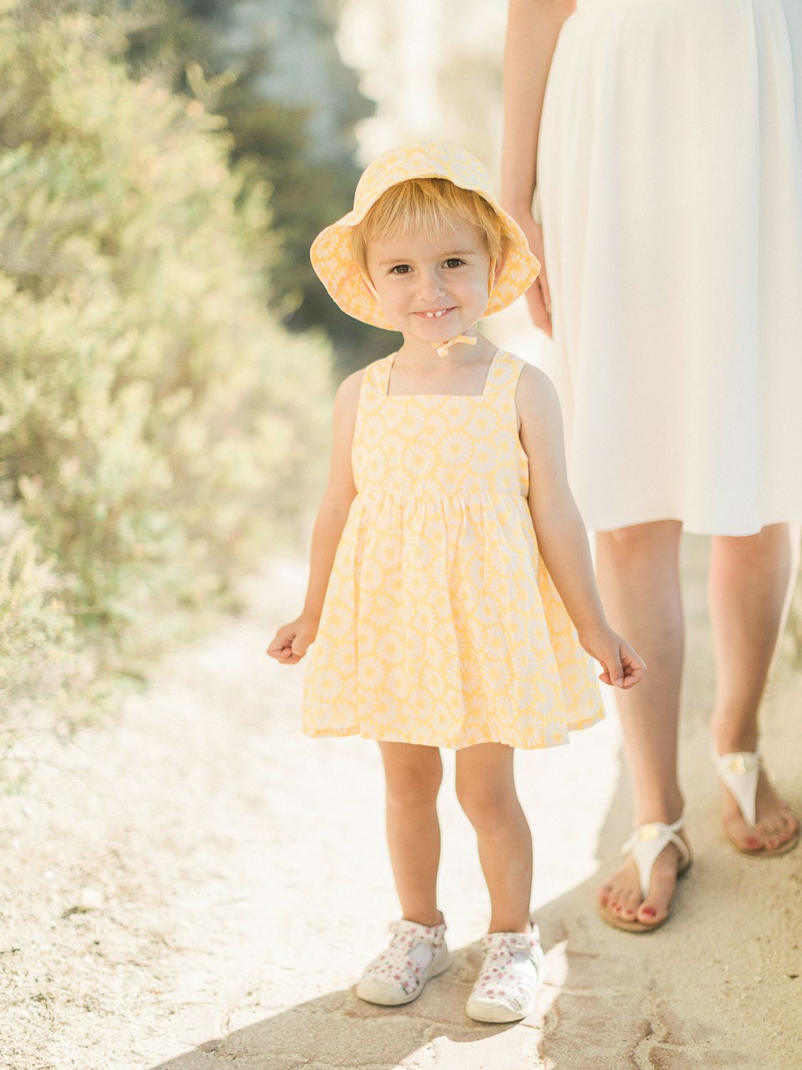 Une petite fille souriante, vêtue d'une robe jaune à fleurs et d'un chapeau de soleil, se tient sur un chemin ensoleillé.