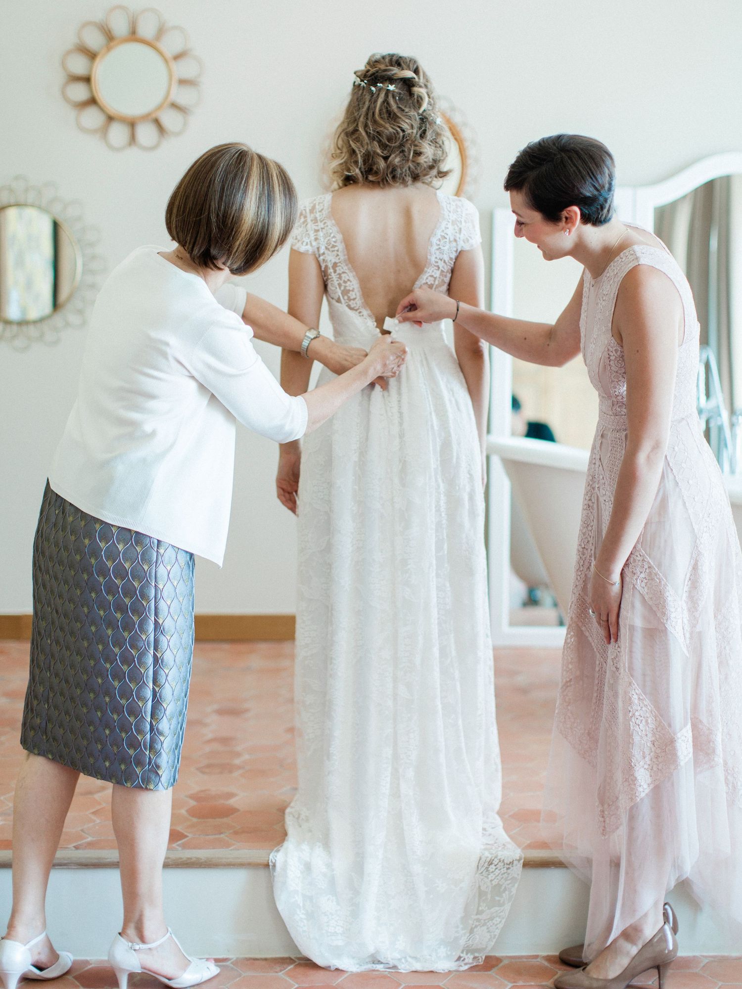 Two women helping a bride fasten her lace wedding dress from behind in an elegant room.