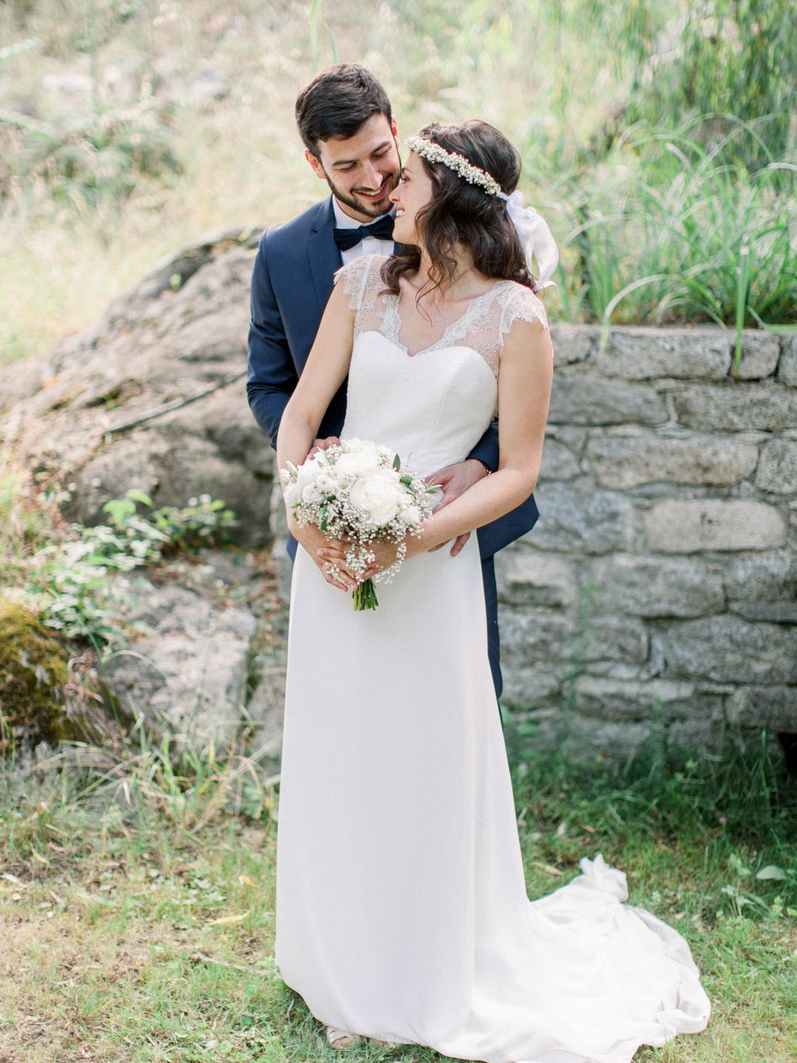 A smiling groom in navy suit embraces bride in white lace gown holding white bouquet outdoors near stone wall.