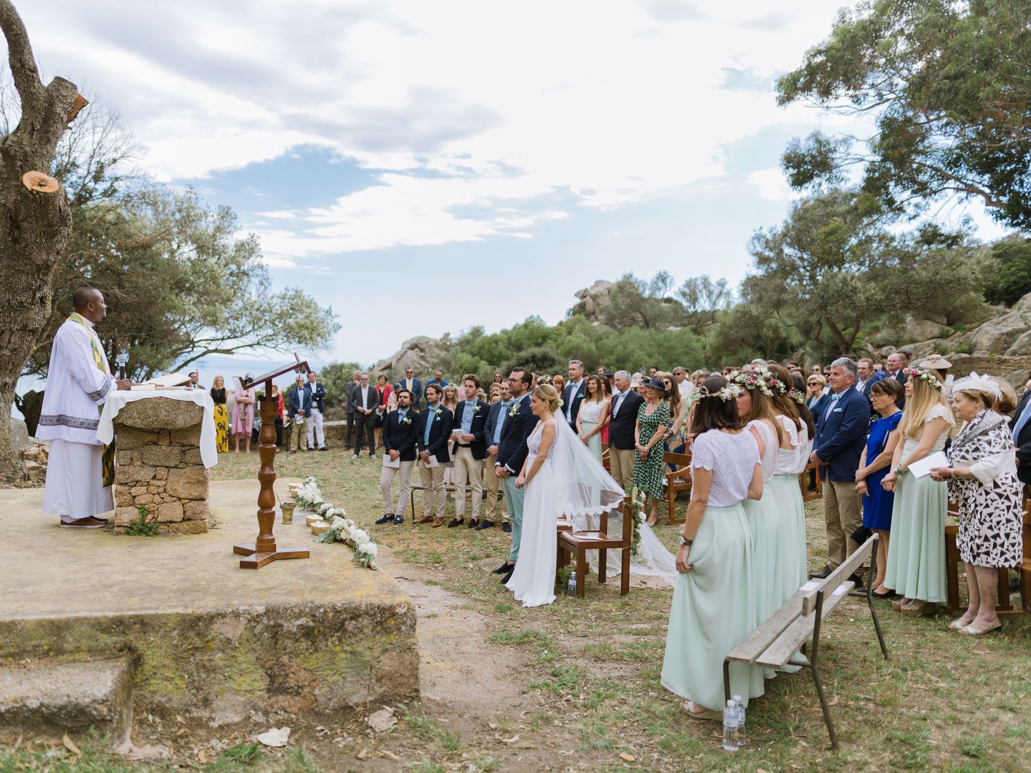 Cérémonie de mariage en plein air, les invités étant rassemblés parmi des ruines antiques et des arbres sous un ciel partiellement nuageux.