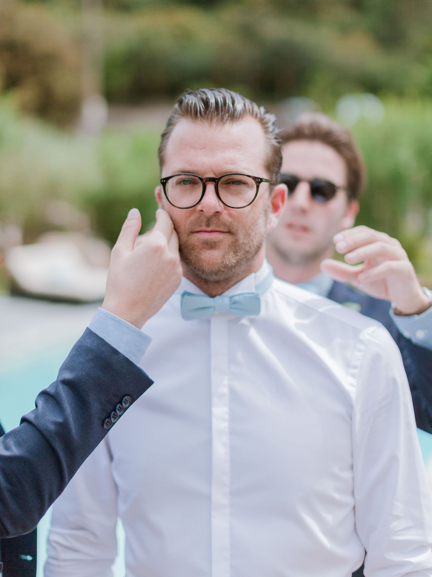 Le marié, en chemise blanche et nœud papillon bleu clair, ajuste ses lunettes à l'extérieur lors de la cérémonie de mariage.