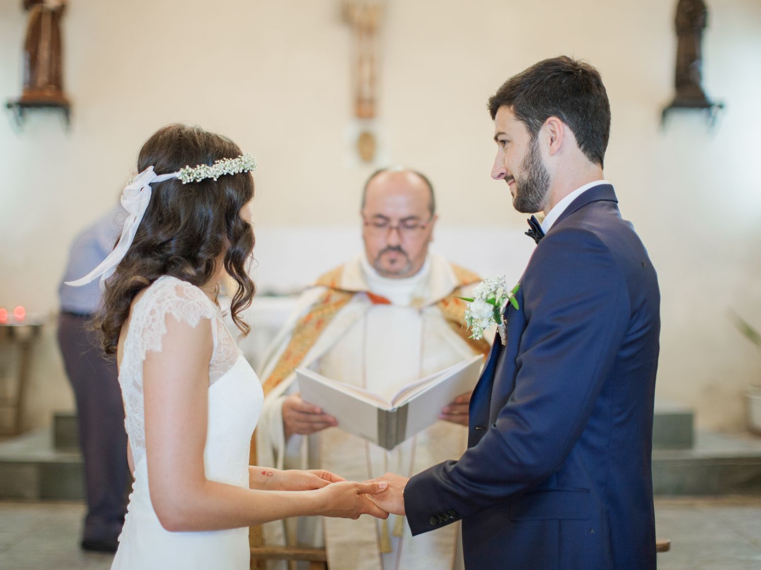 Bride and groom exchange vows at church altar with officiant reading from book during elegant wedding ceremony.