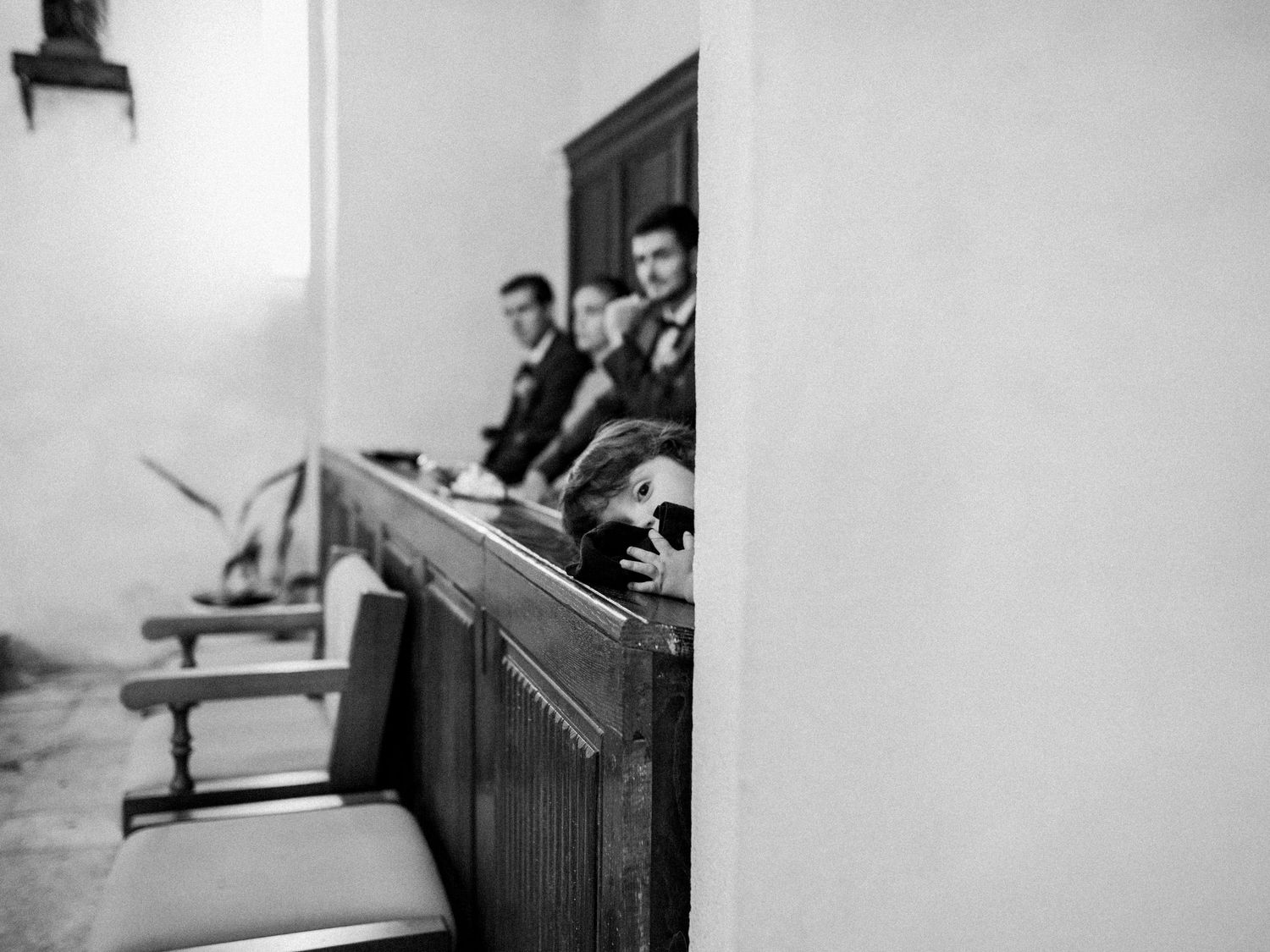 Black and white photo of wedding guests seated in church pews, with a child peeking over the bench.