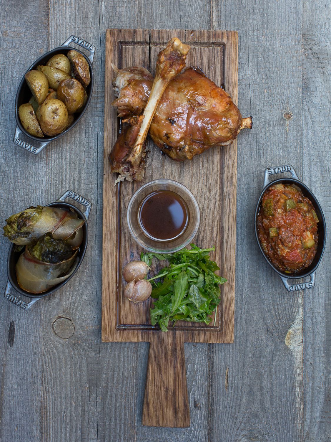 Jarret d'agneau rôti sur planche en bois avec sauce, légumes verts, ail, pommes de terre rôties, aubergines et relish de tomates.